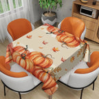 A rectangular table covered with a beige tablecloth printed with large orange pumpkins, scattered autumn leaves, and a small corn motif, shown in a dining room setting with four orange-and-white chairs.