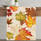 A square fabric placemat with a watercolor autumn leaf pattern in red, orange, yellow, and green on a cream background, displayed on a wooden table.