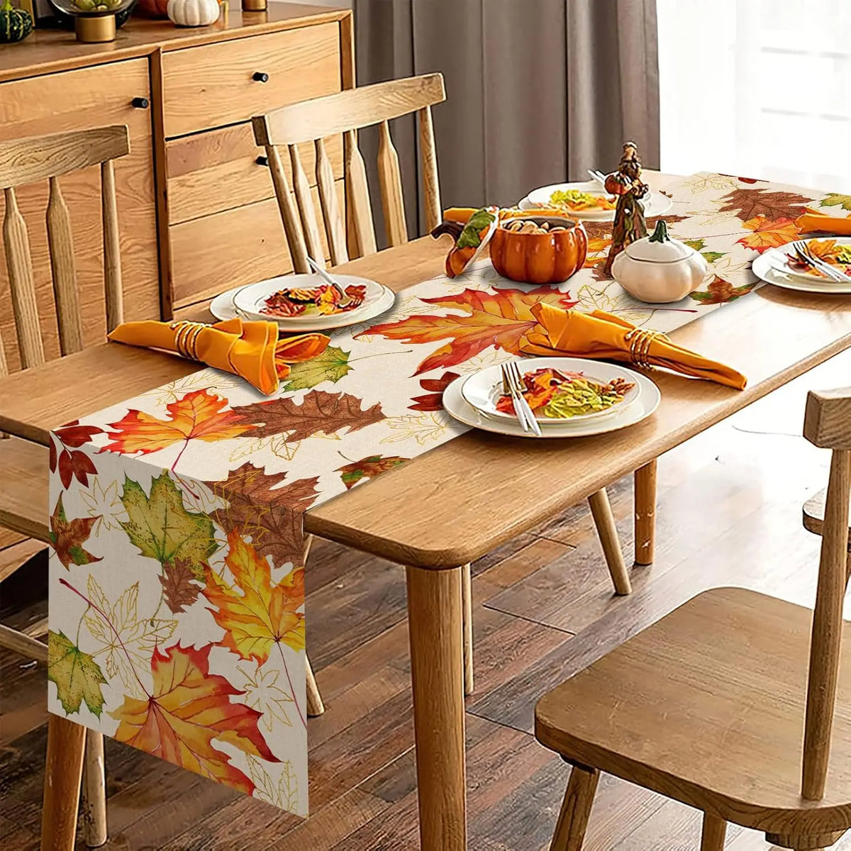 An autumn-themed table runner with colorful fall leaves draped over a wooden dining table, styled with plates, orange napkins, pumpkins, and chairs in a warm dining room setting.