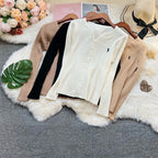 Three ribbed long-sleeve button-front cardigans in cream, black, and beige are arranged on a white faux-fur rug with wooden hangers. The cardigans feature a small embroidered polo player logo on the chest.