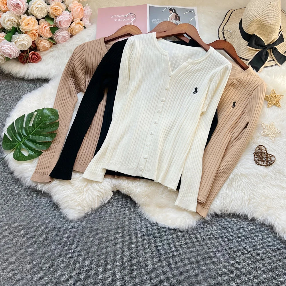 Three ribbed long-sleeve button-front cardigans in cream, black, and beige are arranged on a white faux-fur rug with wooden hangers. The cardigans feature a small embroidered polo player logo on the chest.