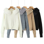 Four women's knit zip-front hooded sweaters with faux-fur or faux-fur-trimmed hoods, displayed on wooden hangers against a white background. The colors shown are off-white, heather gray, tan, and black.