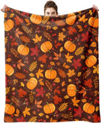 A person holds up a large fleece blanket featuring a fall harvest pattern with orange pumpkins, red and yellow leaves, and pinecones on a dark brown background.